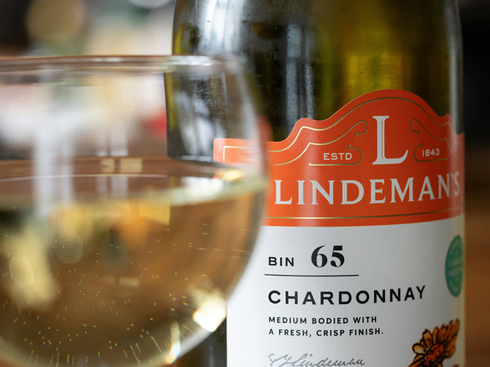 Garrafa de vinho Chardonnay Lindeman’s Bin 65 com taça à frente, destacando o rótulo e a tonalidade clara do vinho branco, símbolo de frescor e sofisticação. 